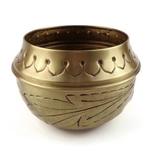 Op de afbeelding: Een ronde messing kom met een metallic gouden afwerking. Het bovenste gedeelte is voorzien van een herhalend schulppatroon, terwijl het onderste gedeelte is versierd met gegraveerde bladachtige motieven. De kom heeft een brede opening.
