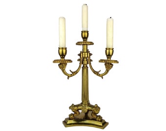 May include: A gold-toned metal candelabra with three candle holders. The candelabra has a decorative base with a scroll design and a central column with ornate details. The candle holders are adorned with decorative accents.