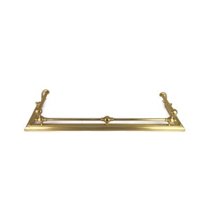 Op de afbeelding: Een gouden metalen schouw met decoratieve eindstukken. De schouw is ontworpen om boven een open haard te worden gemonteerd.