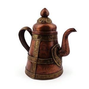 May include: A copper-colored teapot with a decorative design. The teapot has a curved spout, a handle, and a lid with a finial. The body of the teapot is adorned with brass-colored bands and decorative elements. The teapot is likely used for serving tea.