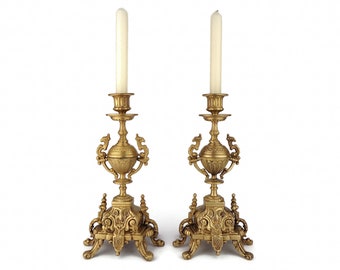 May include: Pair of ornate gold-colored candle holders with white candles. The candle holders have intricate designs and a detailed base. The candles are tall and slender. The background is plain white.