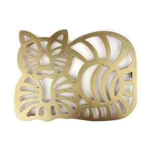 Puede incluir: Un salvamanteles de metal dorado con forma de gato estilizado. El gato está enroscado con la cola envuelta alrededor de su cuerpo. El salvamanteles tiene un diseño recortado que crea un patrón decorativo.