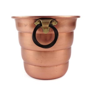Puede incluir: Un cubo de color cobre con un asa de latón y un anillo negro. El cubo tiene un diseño escalonado y una base redondeada. El asa está fijada al borde superior del cubo.