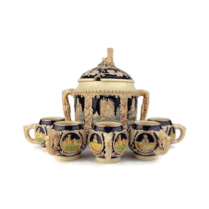 Può includere: Un set di sei tazze in ceramica con un design blu e oro che presenta un castello e dell'uva. Le tazze sono disposte attorno a una brocca in ceramica più grande con un design abbinato.