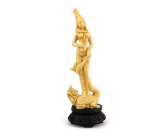 May include: A cream-colored figurine of a woman standing on a turtle. The woman is wearing a headdress and jewelry. The figurine is on a black base.