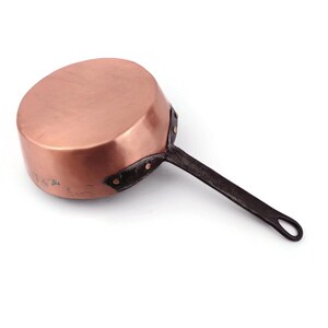 Könnte beinhalten: Ein Kupfertopf mit einem dunklen Metallgriff. Die Pfanne hat einen abgerundeten Körper und ein leicht abgenutztes Aussehen. Der Griff ist mit Nieten befestigt. Küchenutensilien.