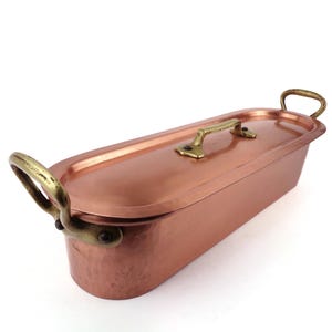 Op de afbeelding: Een koperkleurige, rechthoekige braadpan met een deksel en messing handgrepen. De pan heeft een gehamerde textuur en een gepolijste afwerking. De handgrepen zijn gebogen en aan de zijkanten bevestigd. Het deksel heeft ook een messing handvat.