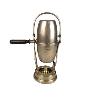 Può includere: Una caffettiera in argento e ottone con un manico nero. La caffettiera è su un supporto con un bruciatore.