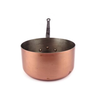 May include: A copper-colored saucepan with a dark metal handle. The pan has a rounded body and a slightly flared rim. The handle is riveted to the pan. The interior of the pan is a darker, aged metal color.