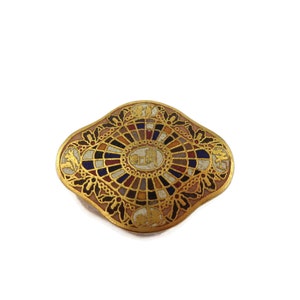 May include: A gold-toned brooch with a colorful mosaic design. The center of the brooch features a circular design with a white background and gold accents. The brooch is surrounded by a decorative border with floral motifs.