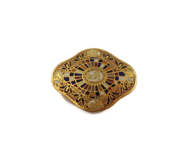 May include: A gold-toned brooch with a colorful mosaic design. The center of the brooch features a circular design with a white background and gold accents. The brooch is surrounded by a decorative border with floral motifs.