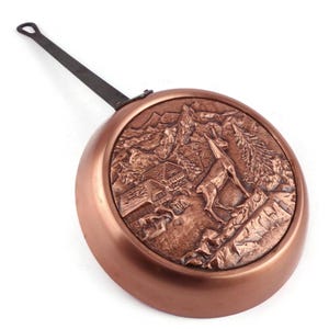 May include: A round copper pan featuring a long, dark metal handle and a detailed embossed relief on its base. The relief depicts a mountain landscape with a chamois standing on rocky terrain, a coniferous tree, and a small wooden cabin structure.