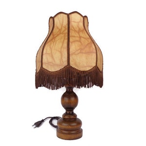 May include: A wooden table lamp with a brown fringed lampshade. The lampshade has a light brown, textured fabric with a pattern of veins.