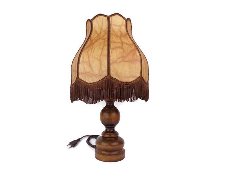 May include: A wooden table lamp with a brown fringed lampshade. The lampshade has a light brown, textured fabric with a pattern of veins.