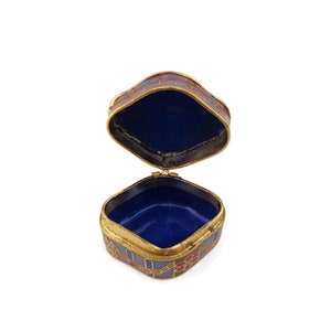 May include: An open, square-shaped trinket box with a blue and red enamel design. The box is made of metal and has a gold trim. The inside of the box is lined with blue velvet.
