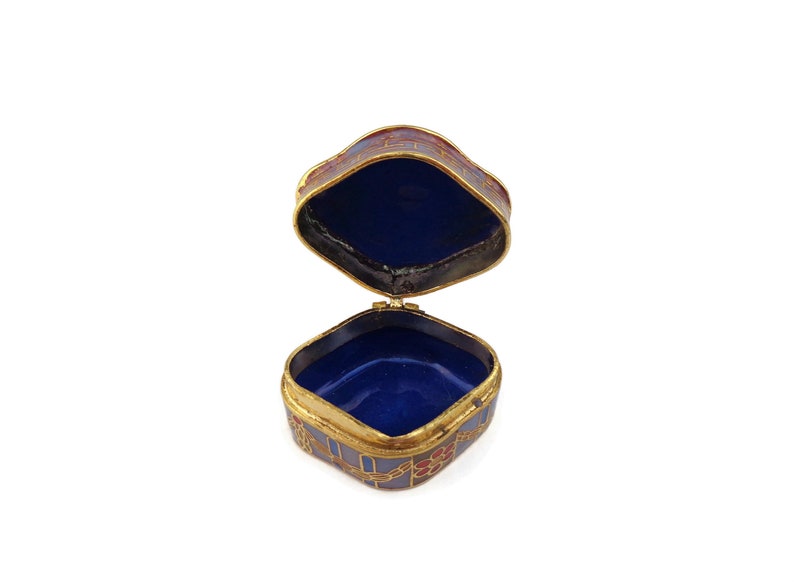May include: An open, square-shaped trinket box with a blue and red enamel design. The box is made of metal and has a gold trim. The inside of the box is lined with blue velvet.