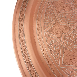 May include: A copper serving tray with an intricate, silver-toned, geometric design. The tray has a raised rim and a smooth, polished surface.