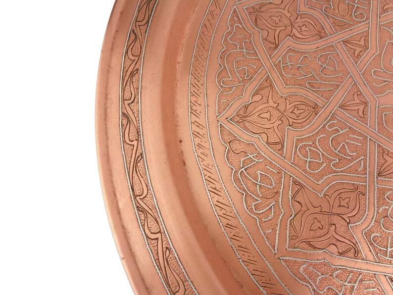 May include: A copper serving tray with an intricate, silver-toned, geometric design. The tray has a raised rim and a smooth, polished surface.