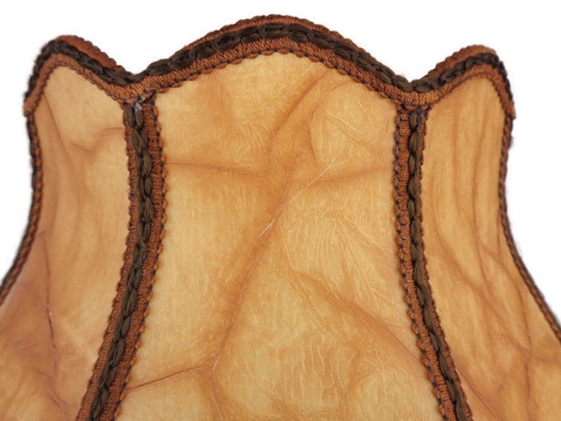 May include: A tan lampshade with a brown scalloped trim. The lampshade has a textured surface and is made of a fabric material.