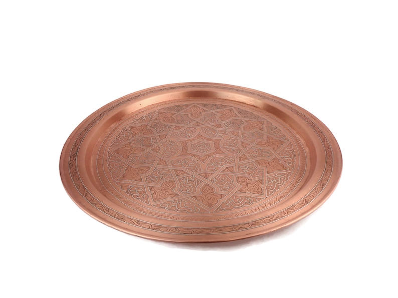 May include: A round copper serving tray with an intricate engraved design. The tray has a raised rim and a polished finish.
