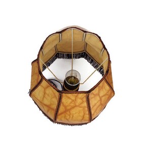 May include: A brown lampshade with a fringe trim and a light bulb inside. The lampshade is made of a textured material that resembles leather.
