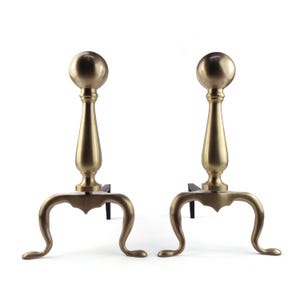 May include: A pair of brass fireplace andirons featuring a tall, slender central column topped with a spherical finial. Each andiron stands on three curved, cabriole-style legs that extend outwards, providing a stable base for the metal structure.