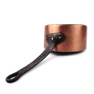 May include: A round copper saucepan with a smooth, metallic finish and a long, dark, textured iron handle. The handle is securely attached to the side of the pan with a metal plate featuring three small, visible copper rivets in a horizontal row.