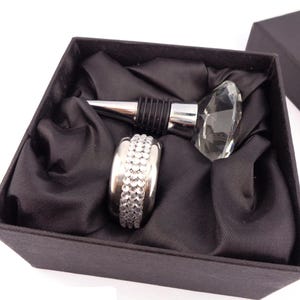 Pode incluir: Uma caixa de presente de veludo preto com um rolha de garrafa prateada e um anel prateado com strass. A rolha de garrafa tem uma parte superior de cristal transparente.
