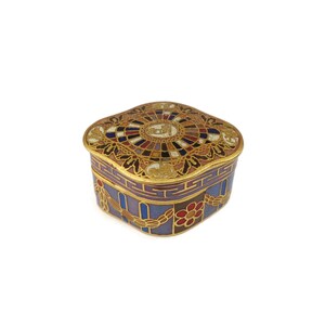 May include: A small, square, decorative box with a gold lid and a blue, red, and gold patterned base. The lid features a circular design with a gold and white center.