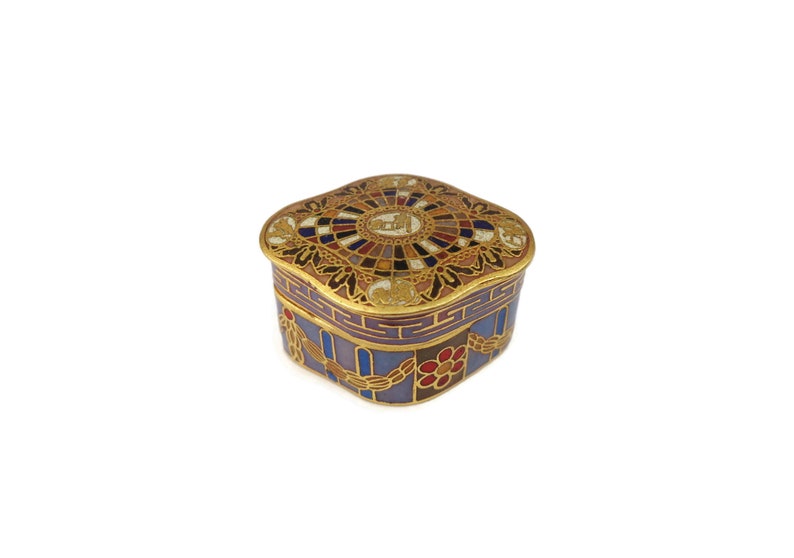 May include: A small, square, decorative box with a gold lid and a blue, red, and gold patterned base. The lid features a circular design with a gold and white center.