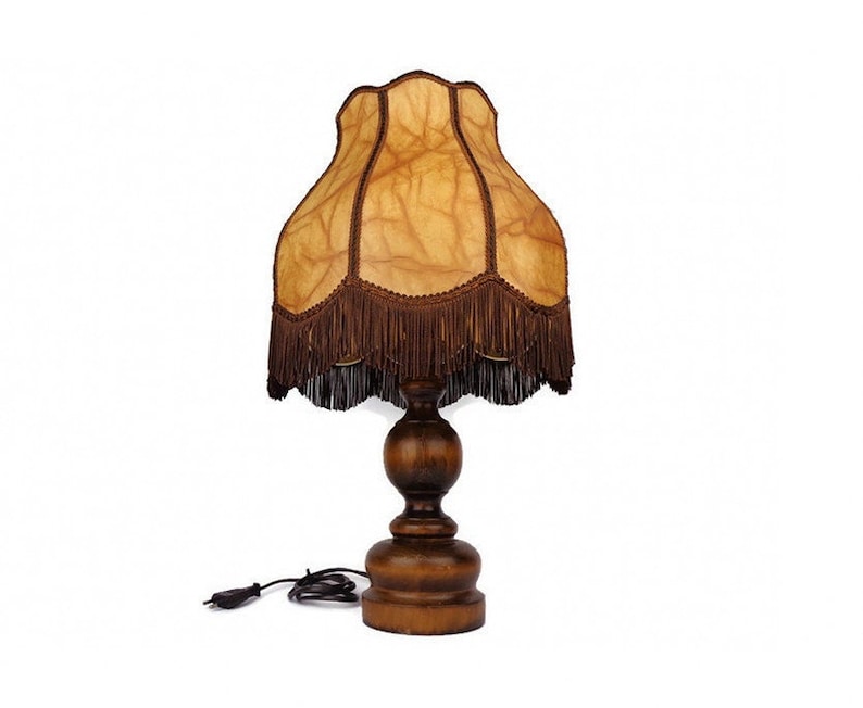 May include: A brown wooden table lamp with a fringed lampshade. The lampshade is made of a light brown fabric with a pattern of veins. The lamp has a round base and a cylindrical stem.