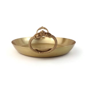 May include: A gold-colored, shallow serving tray with two ornate, ring-shaped handles. The handles feature detailed, textured designs. The tray has a simple, round shape and is made of a shiny, metallic material. The tray is set against a white background.