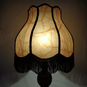 May include: A vintage-style table lamp with a cream-colored, pleated lampshade with black fringe. The lampshade has a textured, crinkled appearance and is lit from within.