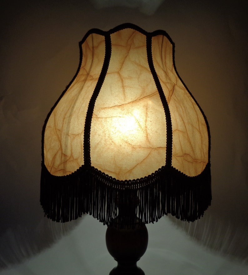 May include: A vintage-style table lamp with a cream-colored, pleated lampshade with black fringe. The lampshade has a textured, crinkled appearance and is lit from within.