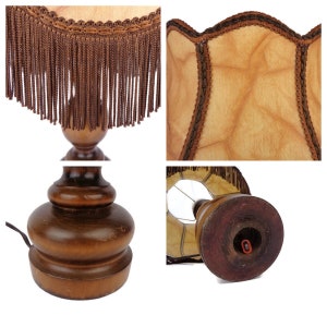 May include: A wooden table lamp with a brown fringed lampshade. The lampshade is made of a light brown fabric with a textured pattern. The lamp base is a dark brown wood with a rounded shape.