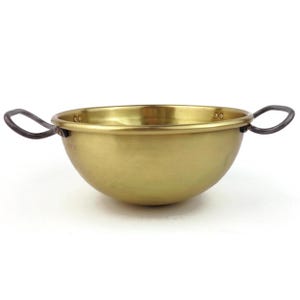 Puede incluir: Un cuenco de color latón con dos asas de metal oscuro. El cuenco tiene forma redondeada y una superficie lisa y reflectante. Las asas son curvas y están unidas a los lados del cuenco. El cuenco probablemente se utiliza para servir o cocinar.