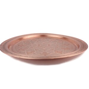 May include: A round copper serving tray with an intricate engraved design. The tray has a raised rim and a polished finish.