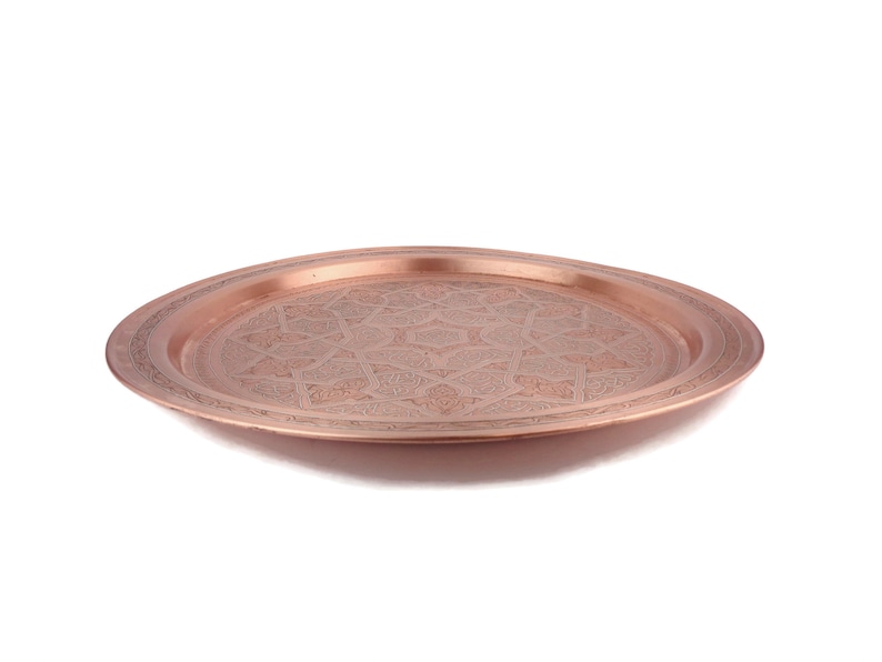 May include: A round copper serving tray with an intricate engraved design. The tray has a raised rim and a polished finish.