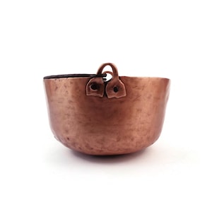 May include: A small, antique copper bowl with a rounded base and two loop handles. The bowl has a dark interior and a hammered texture, showcasing a warm, reddish-brown hue. The handles are attached with rivets.