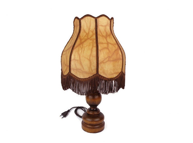 May include: A brown wooden table lamp with a fringed lampshade. The lampshade is made of a light brown fabric with a textured pattern. The lamp has a cord and plug.