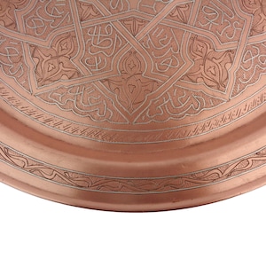 May include: A copper tray with an intricate engraved design. The tray has a circular shape and a raised rim. The design features a repeating pattern of geometric shapes and floral motifs.