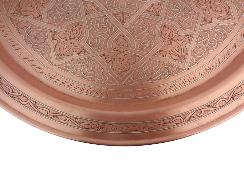 May include: A copper tray with an intricate engraved design. The tray has a circular shape and a raised rim. The design features a repeating pattern of geometric shapes and floral motifs.