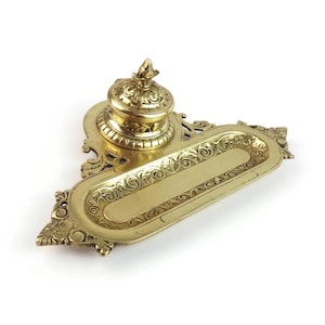 Pode incluir: Um tinteiro de latão polido com um tabuleiro retangular alongado, apresentando volutas ornamentadas e detalhes florais. O recipiente de tinta circular incluído possui uma tampa a condizer com um remate decorativo em latão.