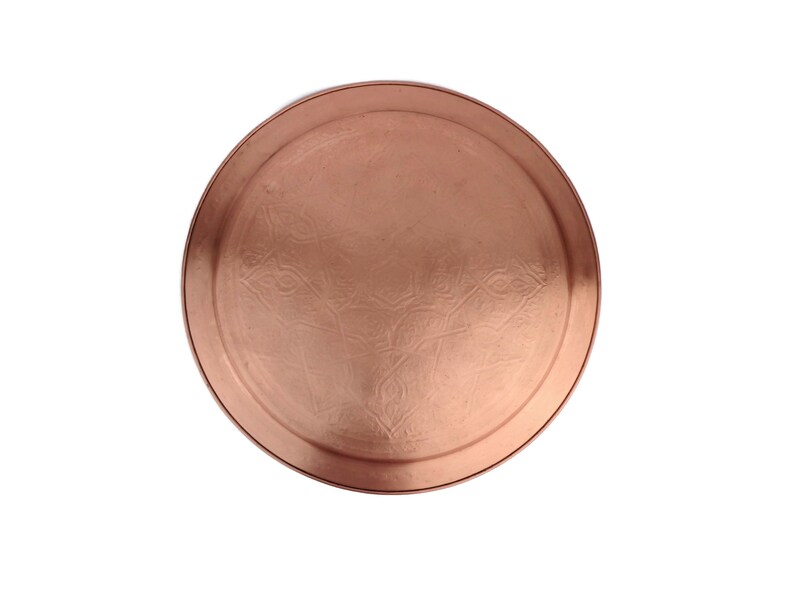 May include: A round copper serving tray with an embossed design. The tray has a smooth, polished surface and a slightly raised rim.