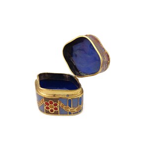 May include: A small, square, decorative box with a hinged lid. The box is made of metal and has a blue, red, and gold enamel design. The inside of the box is lined with blue enamel.