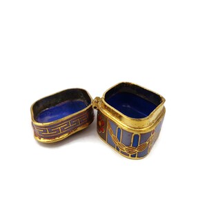 May include: A small, square, gold-colored trinket box with a blue and purple enamel design. The box is open, revealing a blue interior.