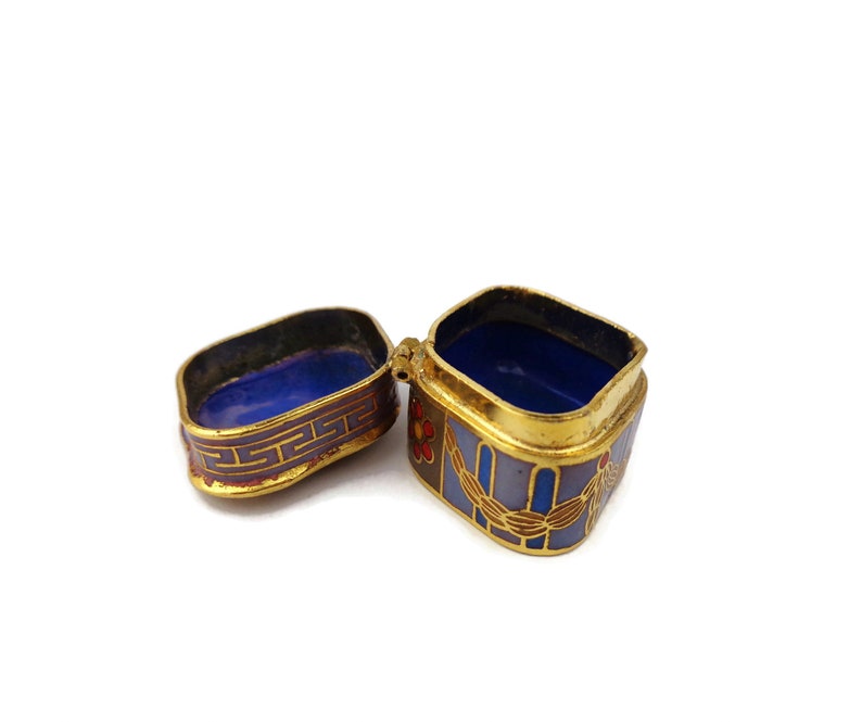 May include: A small, square, gold-colored trinket box with a blue and purple enamel design. The box is open, revealing a blue interior.
