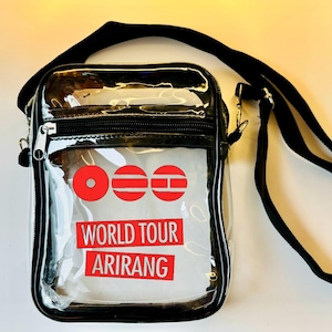 May include: Clear crossbody bag with a black trim and strap. The bag features a red logo and the words "WORLD TOUR ARIRANG" in red block letters. The bag has a zipper closure and a black adjustable strap.