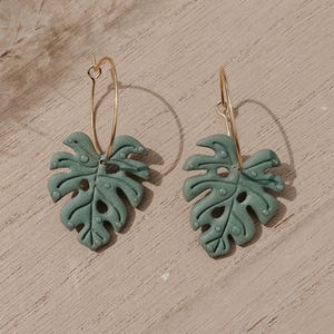 May include: A pair of green Monstera leaf earrings with gold-colored hoop closures. The earrings are a muted green color and feature the distinctive holes and leaf shape of the Monstera plant. The earrings are displayed on a light brown surface.
