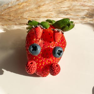 May include: A whimsical arrangement featuring a large, ripe red strawberry adorned with blueberries for eyes, miniature strawberries, and raspberries. Green leaves and stems top the strawberry, creating a playful, fruit-themed design. The background is a soft, neutral color.